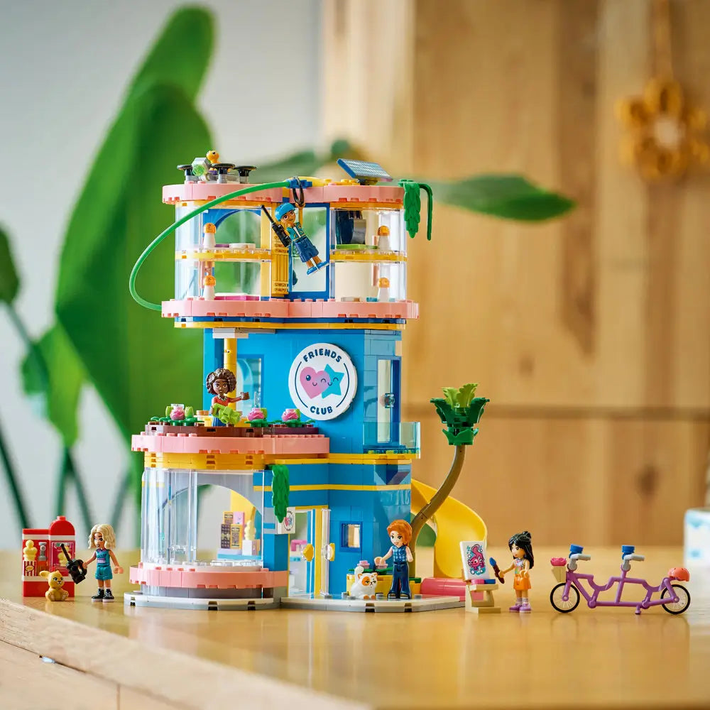 LEGO Friends Heartlake City Friends Club House displayed model shows tall blue and pink plastic building with rounded floors, slide, greenery, mini dolls, bicycle, and café accessories arranged in a bright indoor setting.