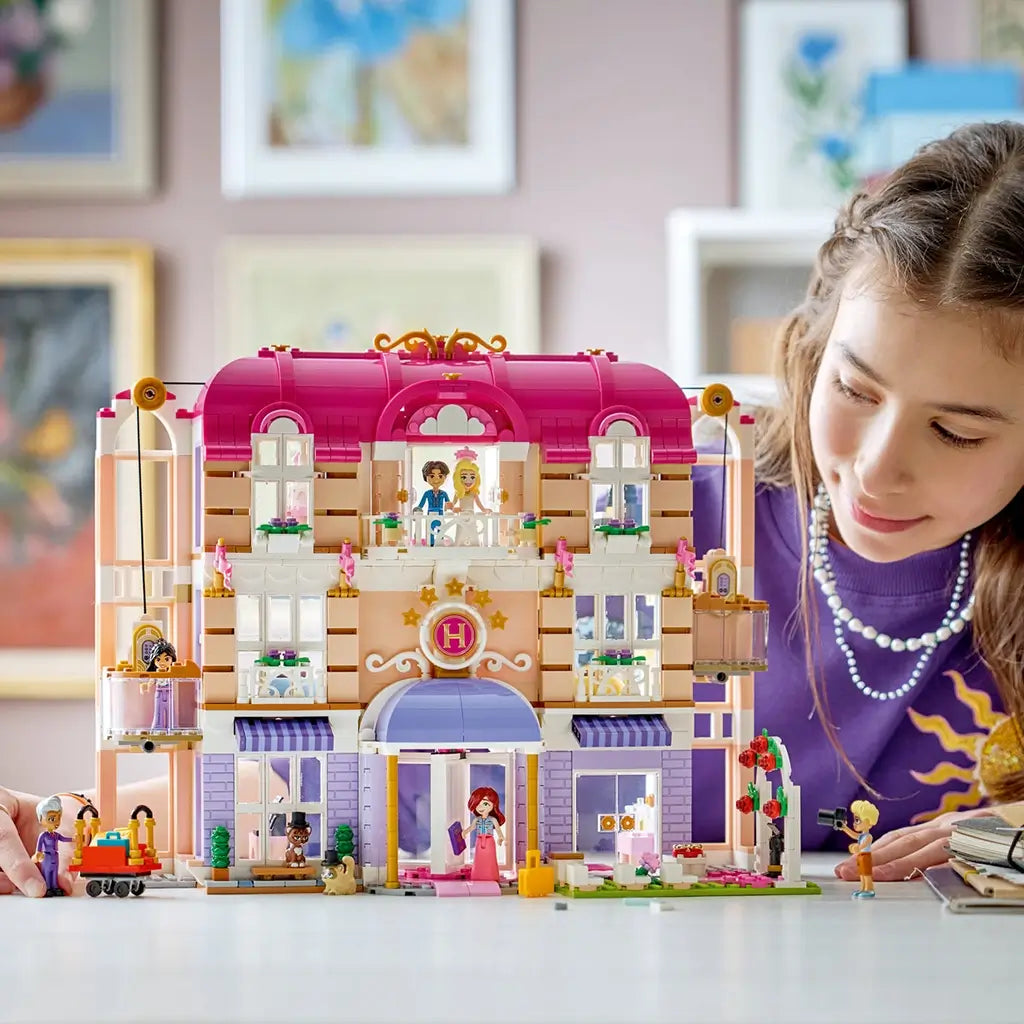 LEGO Friends Heartlake City Grand Hotel play scene shows a child beside the pink, white and lavender hotel facade with figures, elevator, luggage cart and wedding display on a table.