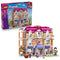 LEGO Friends Heartlake City Grand Hotel set 42704 pairs its retail box with the pink, white and lavender hotel model, featuring balconies, elevator, figures, luggage cart and wedding arch.