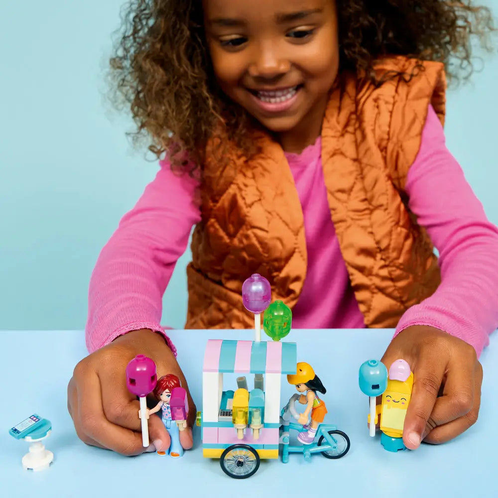 LEGO Friends Ice Cream & Balloon Stand playset used during shared play, pastel bicycle cart with balloons and treats, shown with a child smiling and arranging minidolls and accessories in a bright indoor setting.