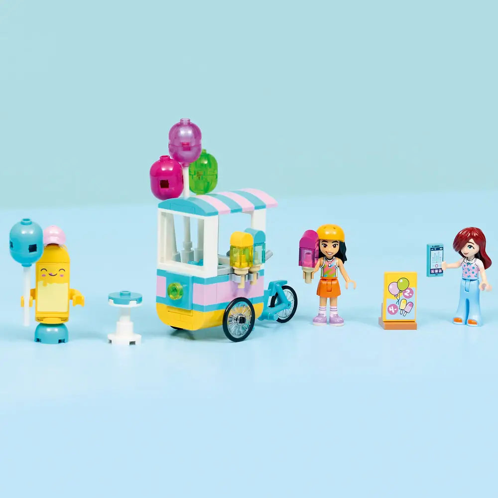 LEGO Friends Ice Cream & Balloon Stand fully assembled, pastel pink and teal bicycle cart with striped canopy, balloon clusters, popsicle holders, and minidolls, neatly displayed on a table against a soft blue background.