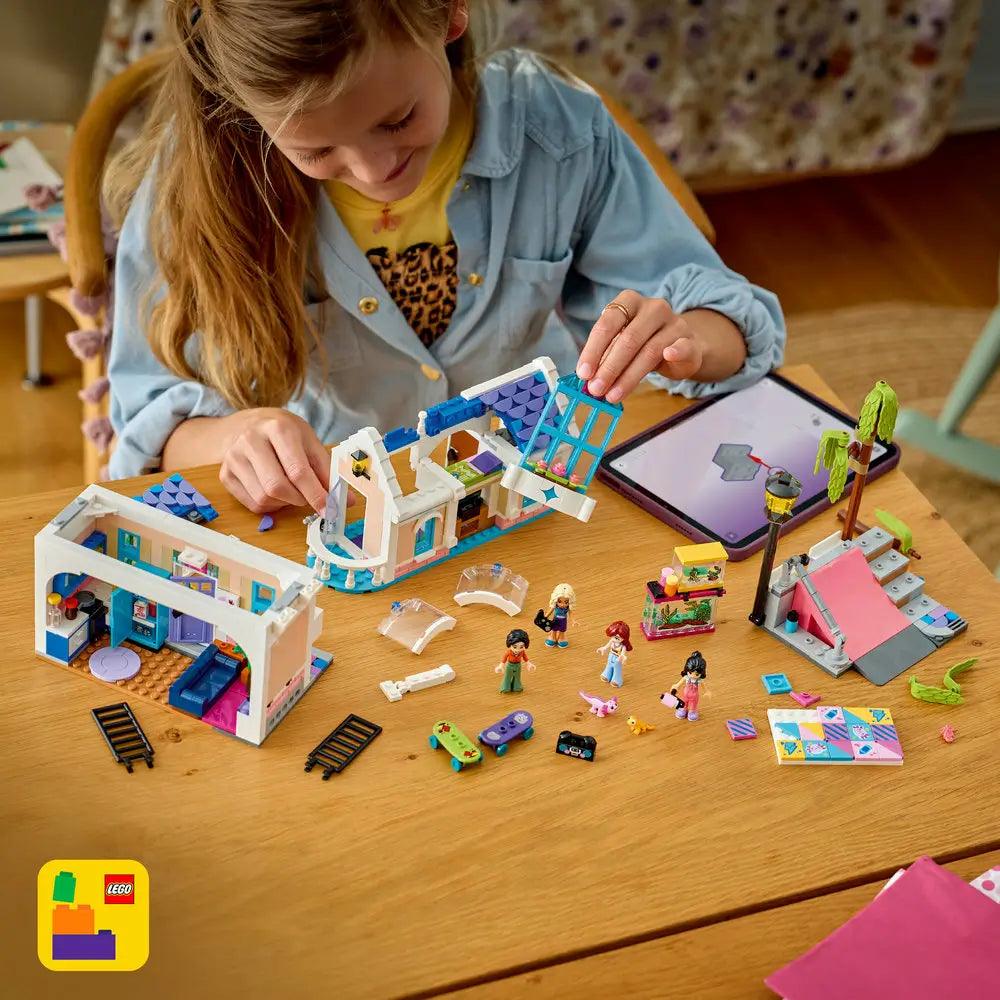 LEGO Friends building toy scene with colorful plastic bricks spread on a wooden table, showing a child assembling a pastel house set with figures, skateboards, accessories, and a tablet used for guided building.