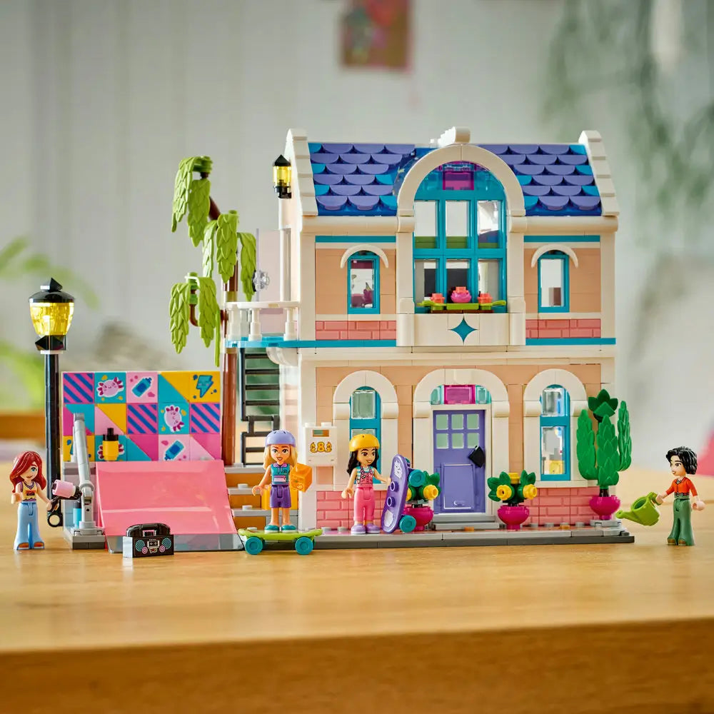 LEGO Friends house display showing pastel peach and pink plastic bricks forming a finished two story home with blue roof, balcony, ramp, trees, planters, and multiple minifigures posed on a tabletop.