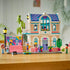 LEGO Friends house display showing pastel peach and pink plastic bricks forming a finished two story home with blue roof, balcony, ramp, trees, planters, and multiple minifigures posed on a tabletop.