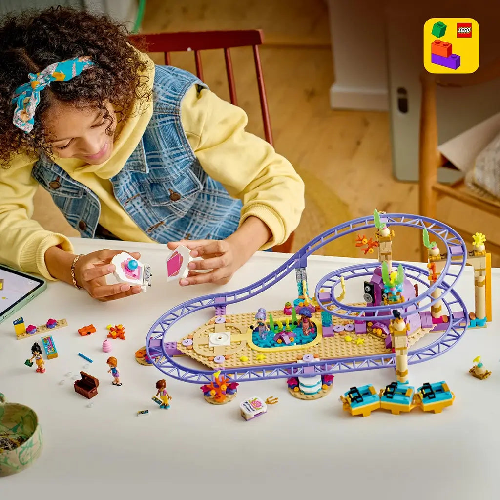 LEGO Friends Mermaid Roller Coaster Ride building scene shows purple plastic tracks and colorful ocean decor as a child assembles accessories beside mini-dolls on a white table.