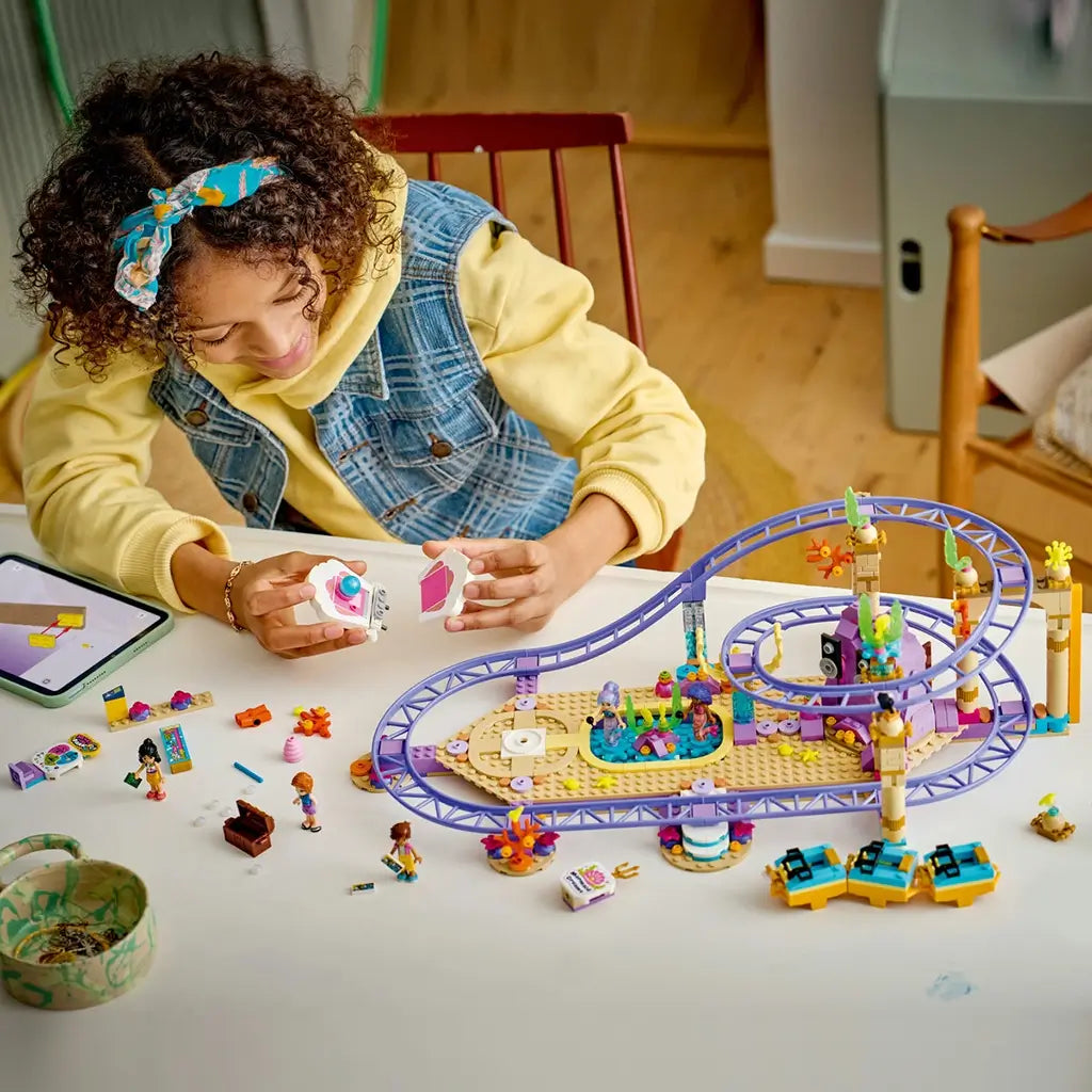 LEGO Friends Mermaid Roller Coaster Ride playset with purple tracks and colorful sea-themed pieces is being assembled by a child at a table with mini-dolls and loose bricks.