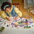 LEGO Friends Mermaid Roller Coaster Ride playset with purple tracks and colorful sea-themed pieces is being assembled by a child at a table with mini-dolls and loose bricks.