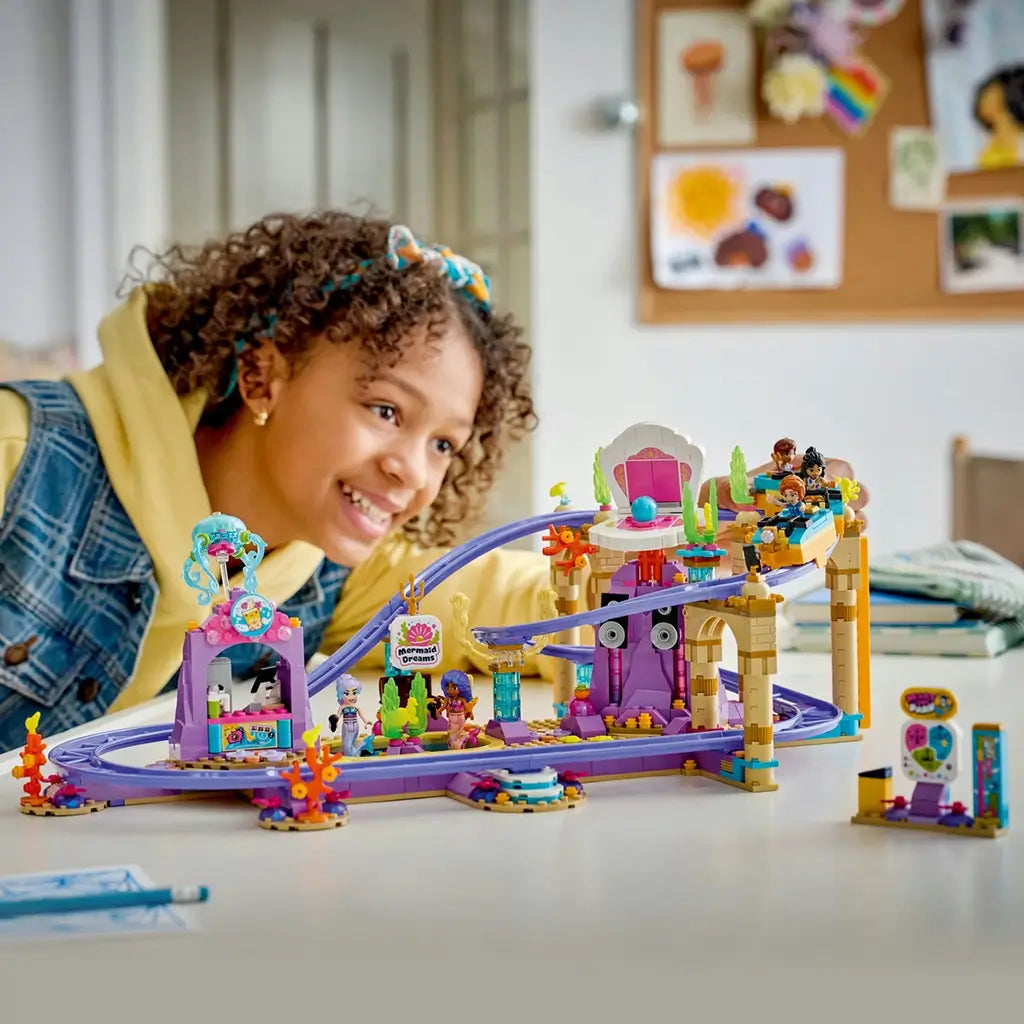 LEGO Friends Mermaid Roller Coaster Ride play scene features purple tracks, colorful ocean-themed structures and a coaster train as a smiling child watches from behind the table.