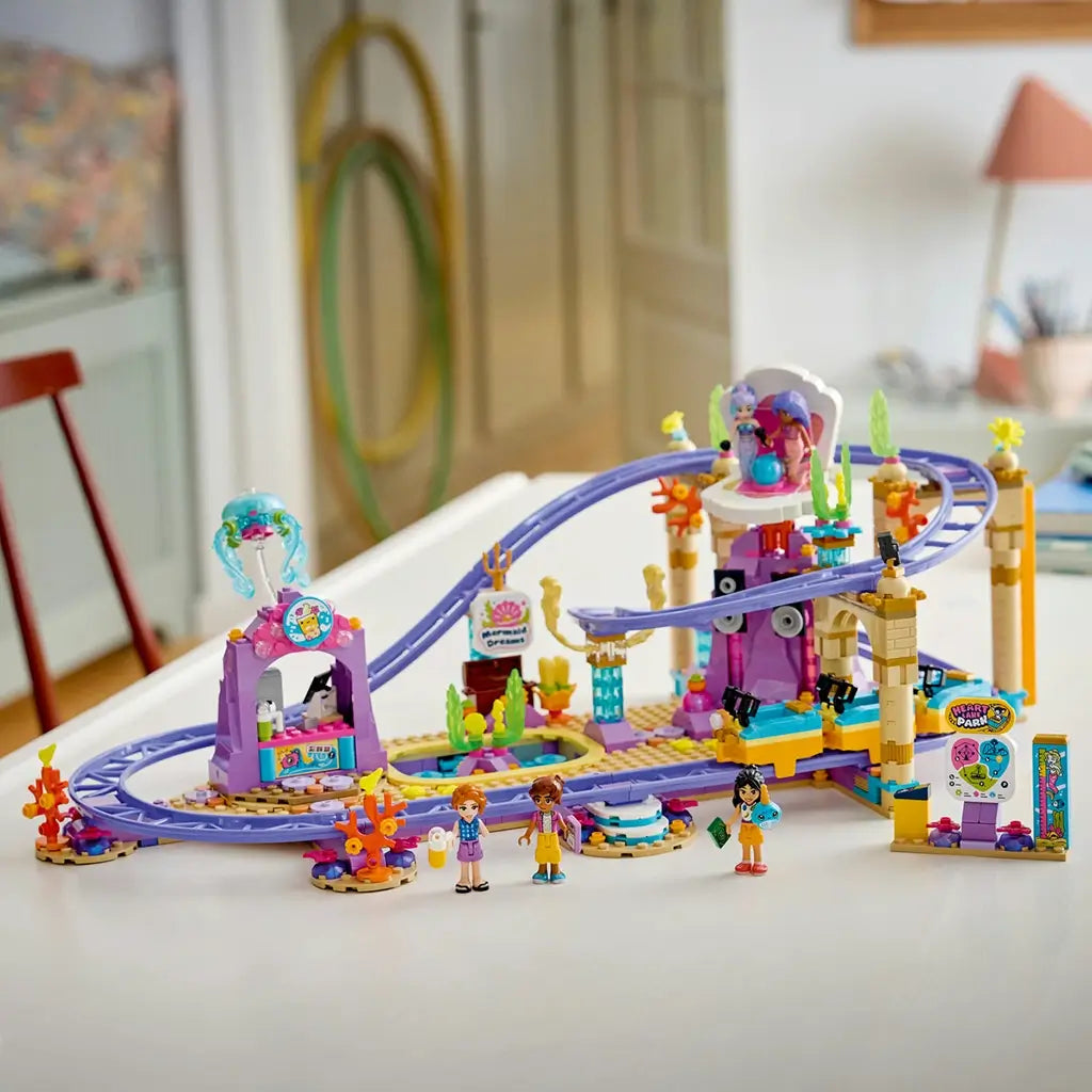LEGO Friends Mermaid Roller Coaster Ride set 42703 features purple plastic tracks, colorful sea-themed scenery, a coaster train and mini-dolls displayed on a white table.