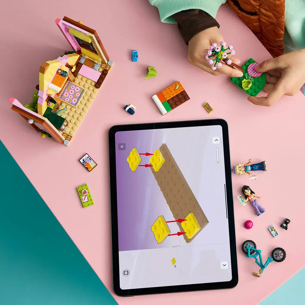 LEGO Friends Nature Glamping Cabin build in pink and brown plastic, with a child assembling flower scenery beside loose pieces and digital instructions on a tablet.
