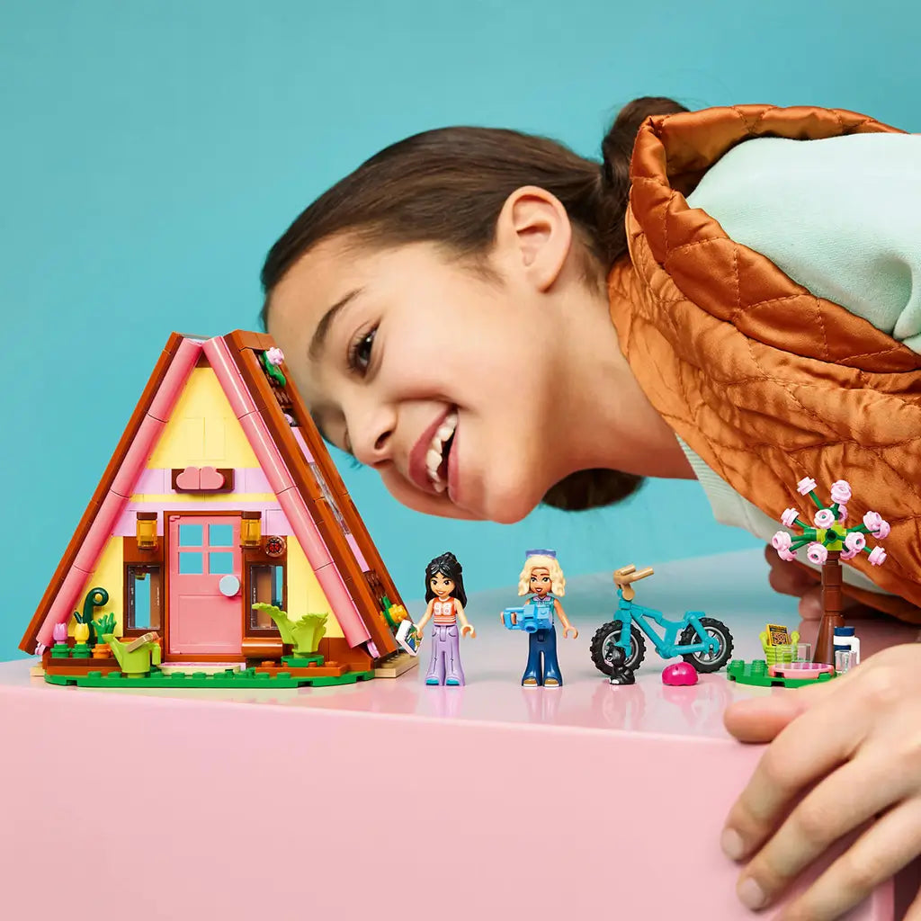 LEGO Friends Nature Glamping Cabin set in pink, yellow and brown plastic, with A-frame cabin, two mini-dolls, teal bike and flower tree as a child plays nearby.