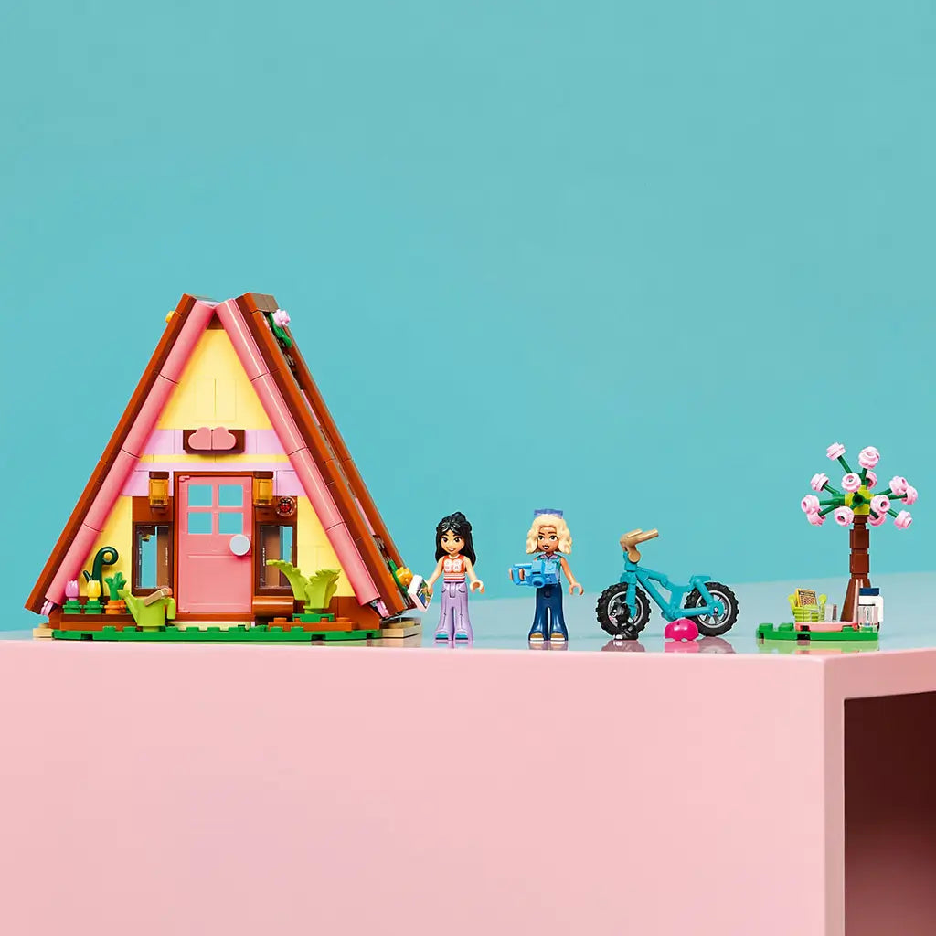 LEGO Friends Nature Glamping Cabin set in pink, yellow and brown plastic, with A-frame cabin, two mini-dolls, teal bike and flower tree displayed on a pink shelf.
