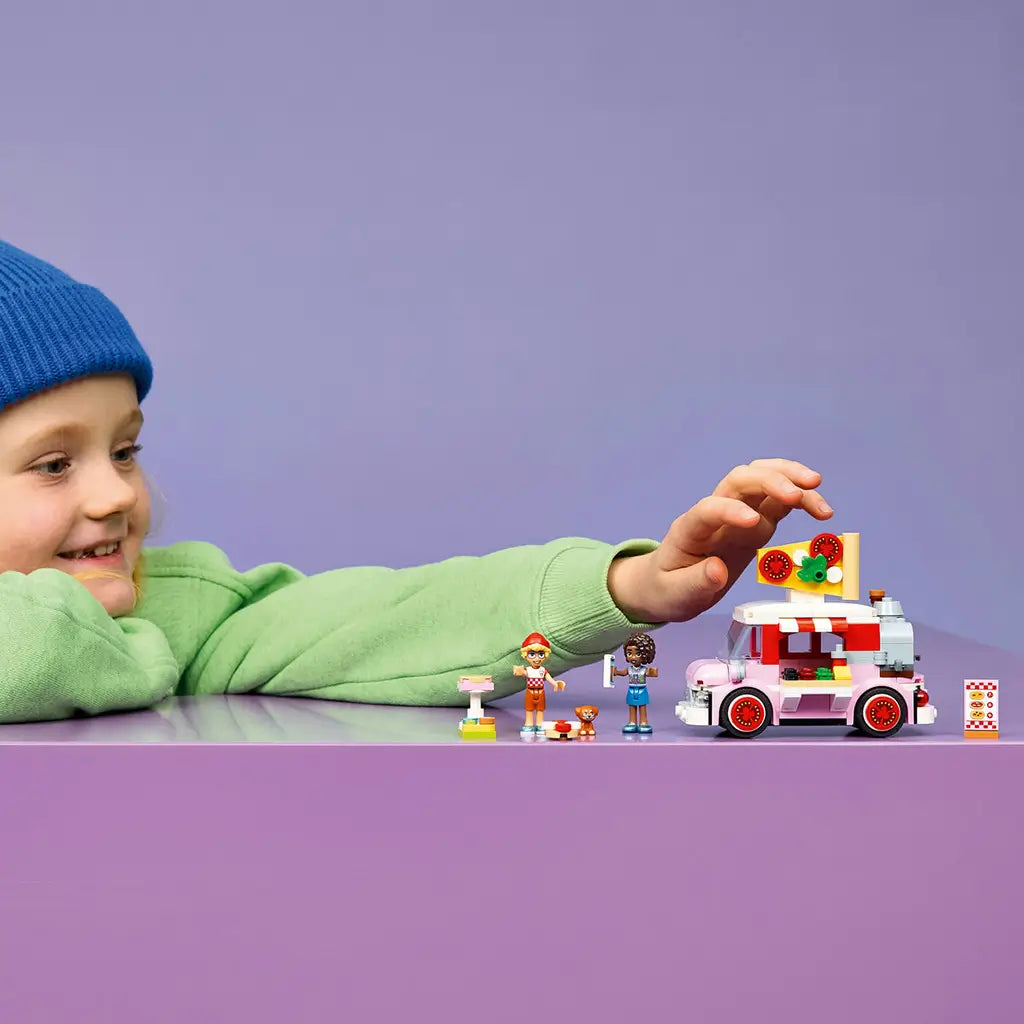 LEGO Friends Pizza Truck set in pink, red, white and gray plastic, displayed with two mini-dolls and accessories as a child reaches toward the model during play.