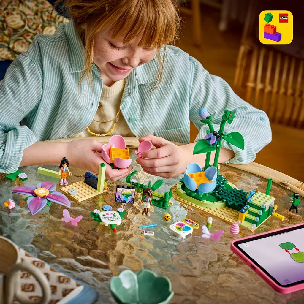 LEGO Friends Spinning Flower & Fairy Teacup Ride building scene features pink, blue and green flower seats as a child assembles the colorful set beside mini-dolls and a tablet.