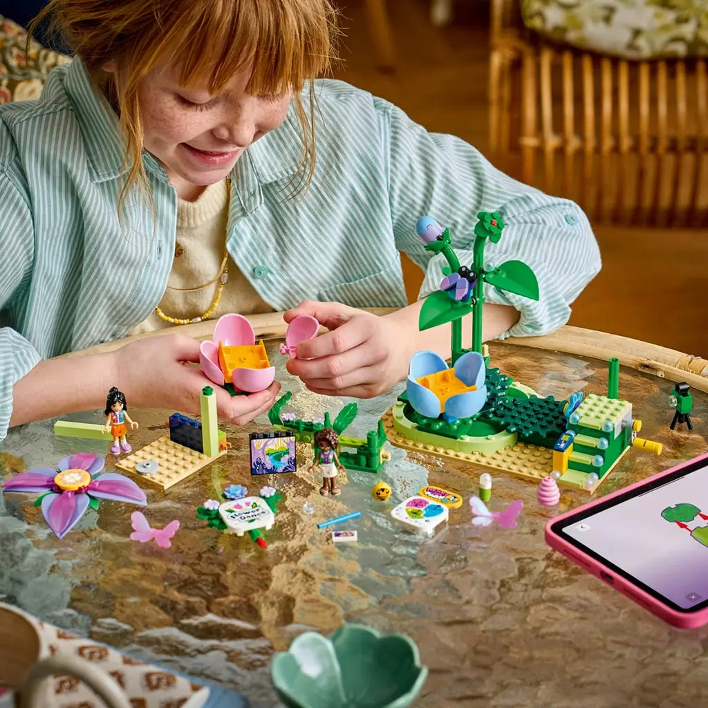 LEGO Friends Spinning Flower & Fairy Teacup Ride building scene shows pink, blue and green plastic pieces as a child assembles flower seats beside mini-dolls and a tablet.