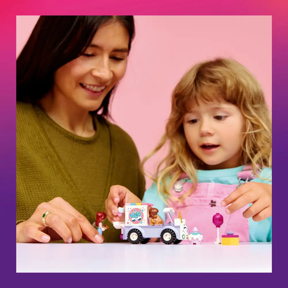 LEGO Friends Unicorn Cake Delivery Car playset shown in use with pastel purple and white plastic pieces as an adult and child sit together, smiling and guiding mini-doll figures and cake accessories during imaginative building play on a pink backdrop.