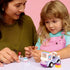LEGO Friends Unicorn Cake Delivery Car lifestyle image shows an adult and child smiling while holding mini-doll figures and pastel cake accessories, with the unicorn delivery truck nearby on a table against a soft pink background.