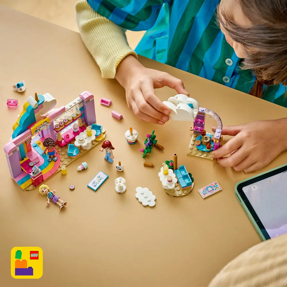 LEGO Friends building toy scene with pastel café pieces spread on a table, showing a child assembling unicorn themed structures, dessert stands, minifigures, and accessories using digital guided instructions.