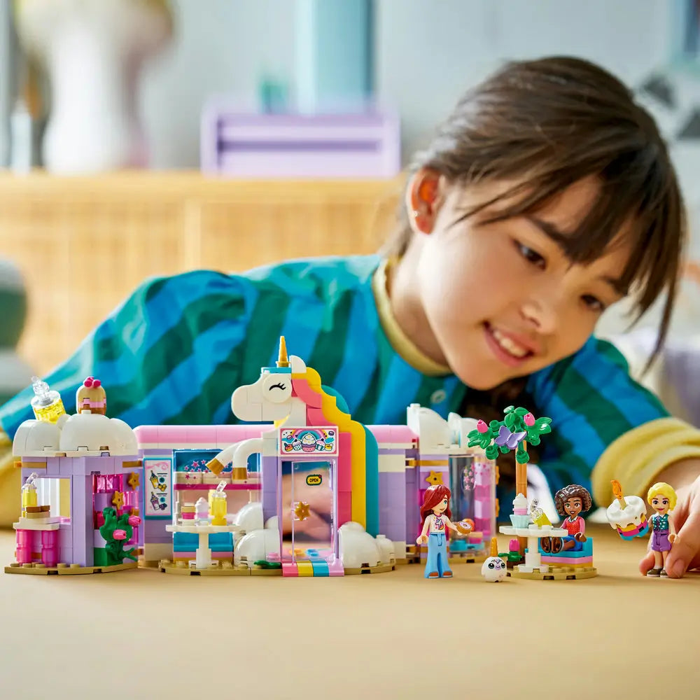 LEGO Friends playset lifestyle scene with a smiling child interacting with a completed unicorn café, moving minifigures between dessert tables, serving areas, and outdoor seating to create fantasy café stories.