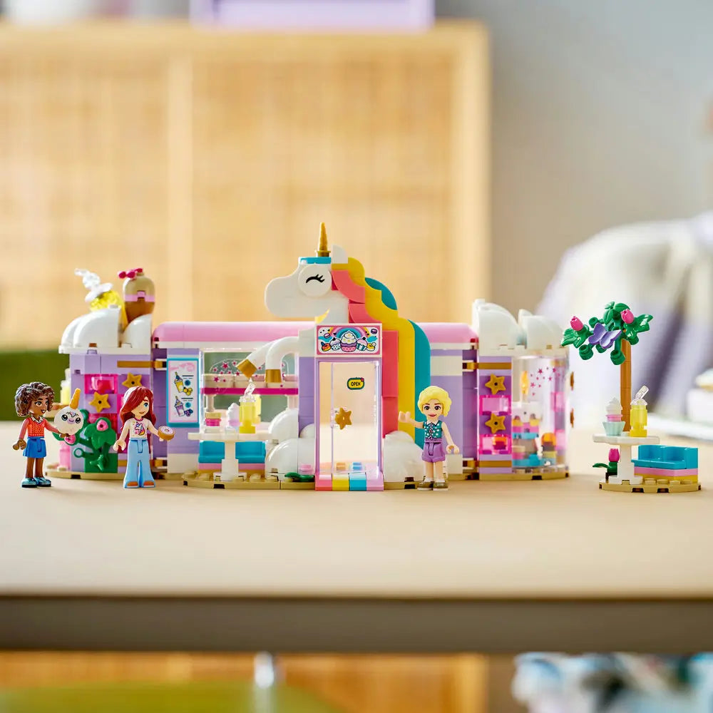 LEGO Friends unicorn café display showing pastel white, pink, and lavender plastic bricks forming a finished café with rainbow doorway, unicorn details, dessert tables, plants, and minifigures posed on a tabletop.
