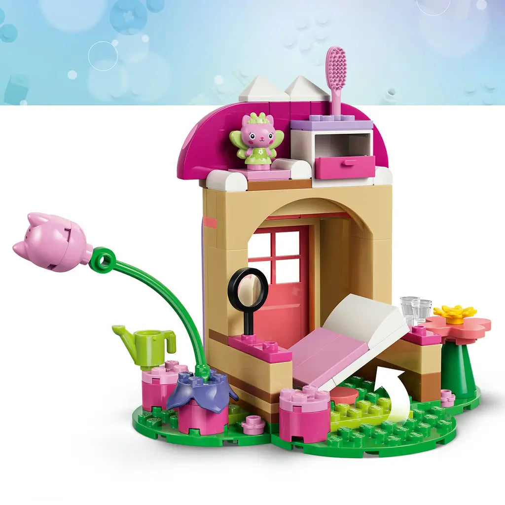 LEGO Gabby's Dollhouse garden house in pink, tan and green plastic bricks, opened to show a bed, drawer, mirror, flower table, watering can and small fairy cat figure.