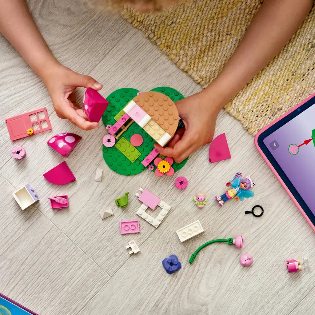 LEGO Gabby's Dollhouse garden house in pink, tan and green plastic bricks is assembled by a child on a light wood floor, with loose pieces, figures and a tablet nearby.