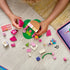 LEGO Gabby's Dollhouse garden house in pink, tan and green plastic bricks is assembled by a child on a light wood floor, with loose pieces, figures and a tablet nearby.