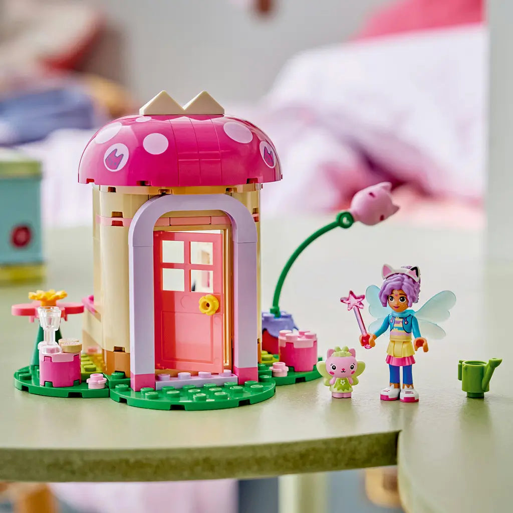 LEGO Gabby's Dollhouse garden house in pink, tan and green plastic bricks, displayed with mushroom roof, pink door, fairy and cat figures, flower stem and watering can.