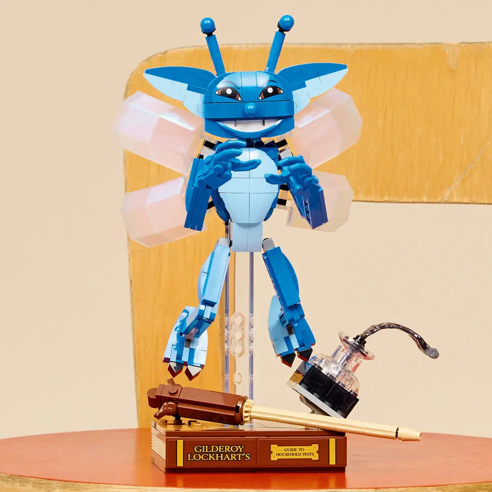 LEGO Cornish Pixie model posed mid-air with translucent wings spread, blue body and antennae visible, displayed on a brown book-themed base with wand and inkwell accessories in a lifestyle setting.
