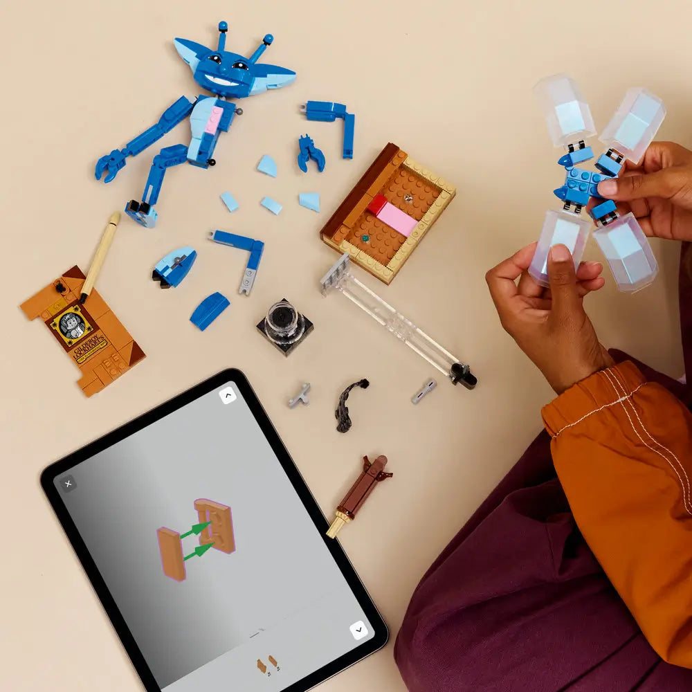 Builder assembling LEGO Cornish Pixie set with blue body parts, translucent wings, book base, and accessories laid out on a table, following digital building instructions on a tablet.