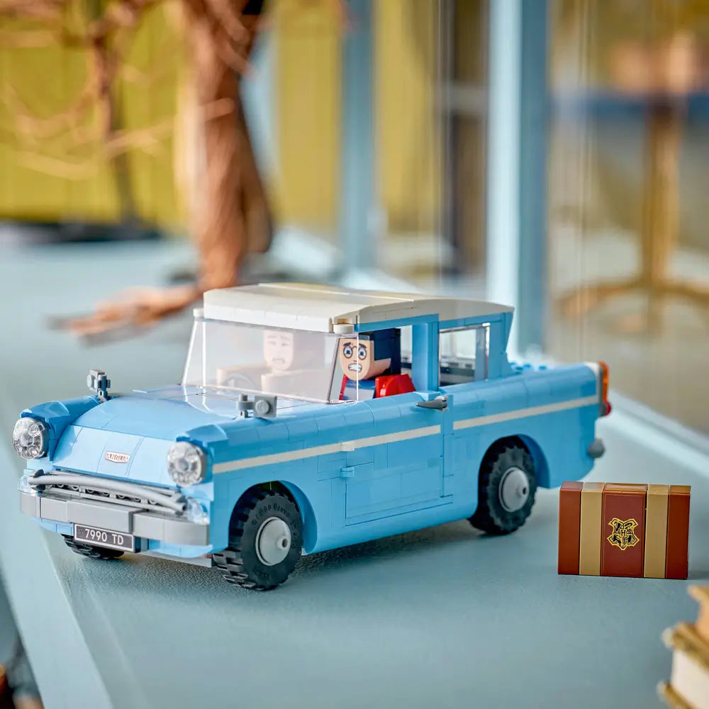 Completed LEGO Harry Potter Enchanted Flying Ford Anglia model in light blue plastic with white roof, black wheels, and minifigures, displayed on a tabletop beside a brown Hogwarts trunk accessory in a play scene.