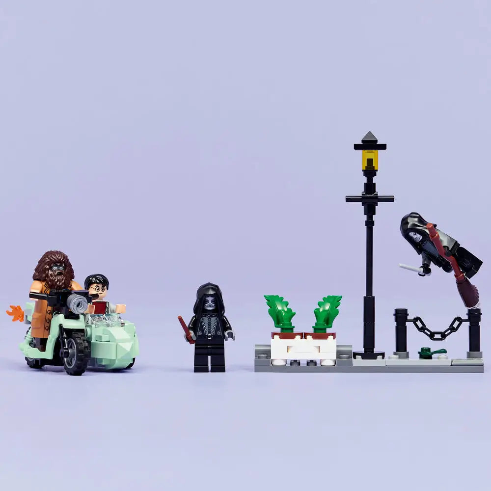 LEGO Harry Potter display scene features multicolor plastic minifigures arranged on a light surface, showing Hagrid and Harry on a motorbike, Death Eaters in pursuit, street lamps, plants, and action poses against a soft backdrop.