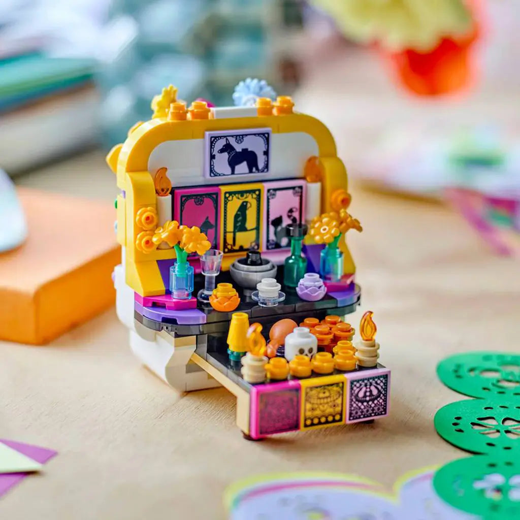 LEGO Altar of the Dead opens into a colorful brick altar with yellow flowers, framed animal art, bottles, candles, food offerings, and a skull on a tabletop.