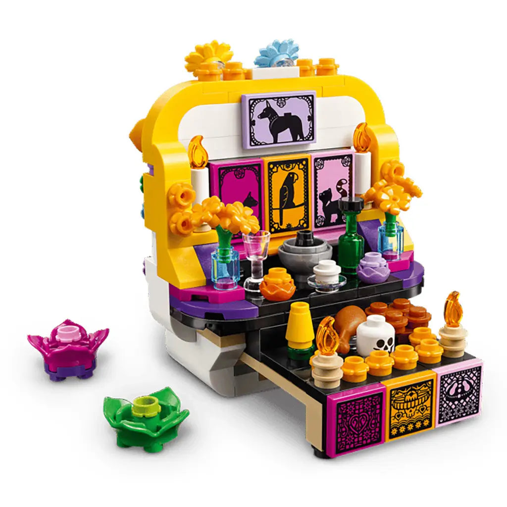 LEGO Altar of the Dead in white, yellow, purple, and black plastic bricks opens to reveal a colorful altar with flowers, bottles, candles, food, and framed animal art.