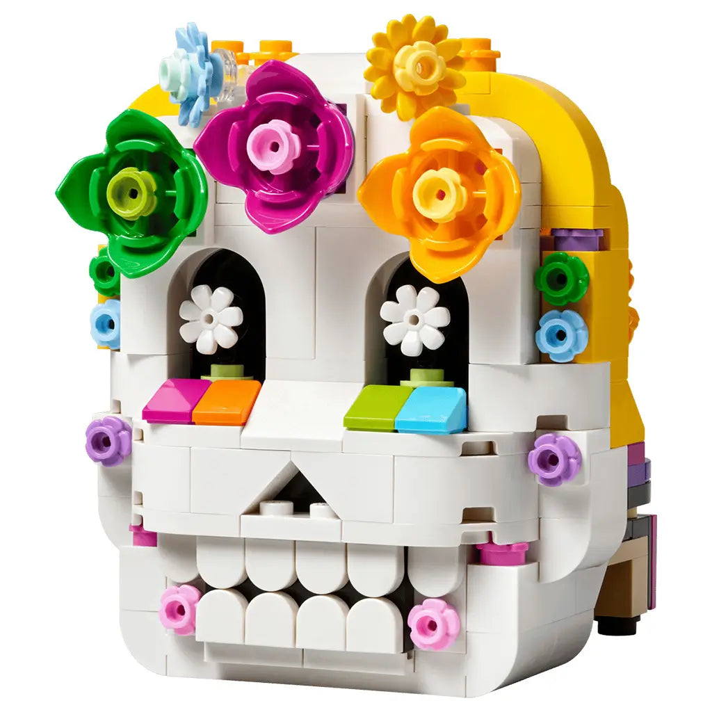 LEGO Altar of the Dead skull in white plastic bricks has a rounded form with black eye sockets, colorful flower decorations, yellow trim, and a toothy grin on white.