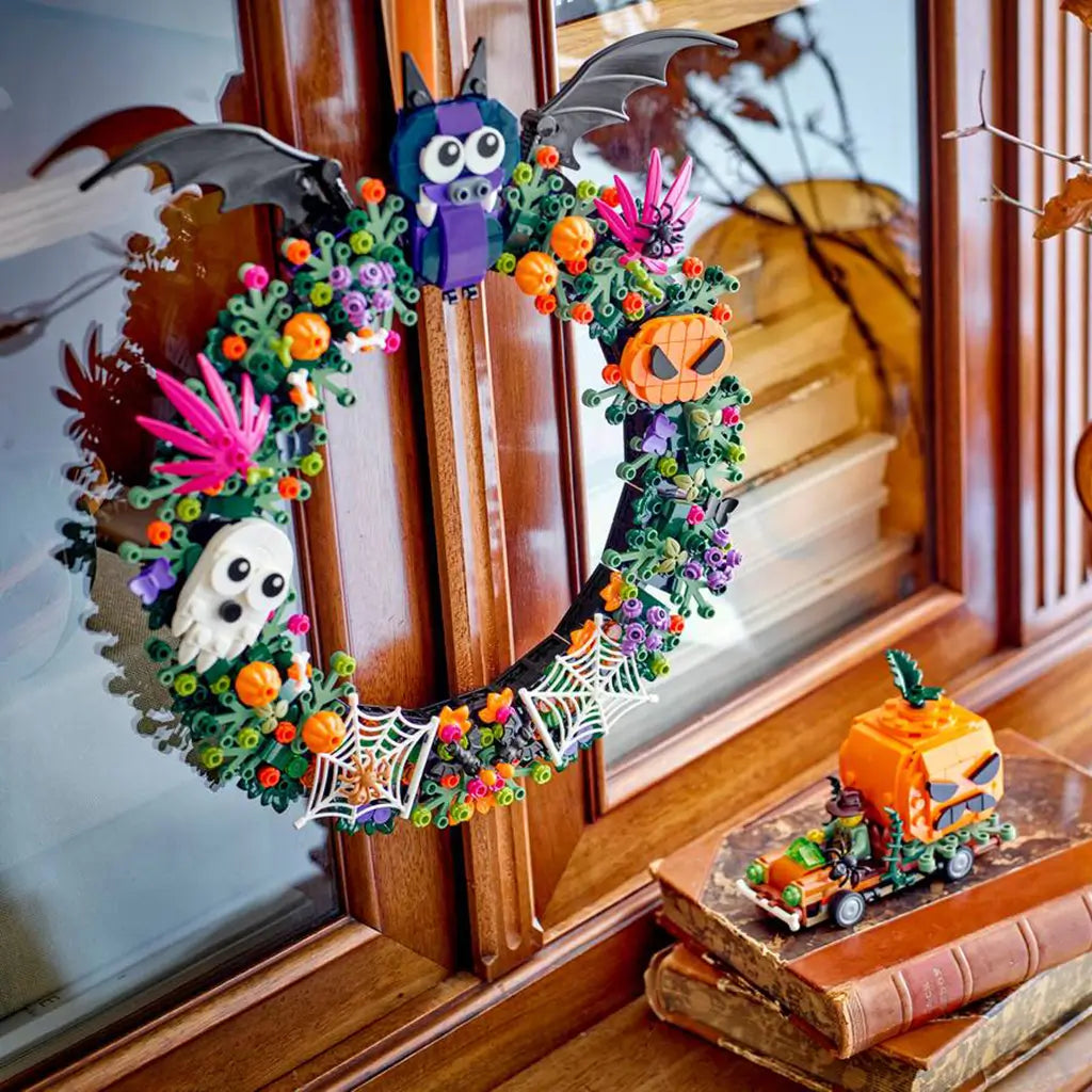 LEGO Halloween wreath in green plastic bricks features orange pumpkins, purple flowers, a white ghost, black bat, spiderwebs, and monster faces displayed on a wooden window.