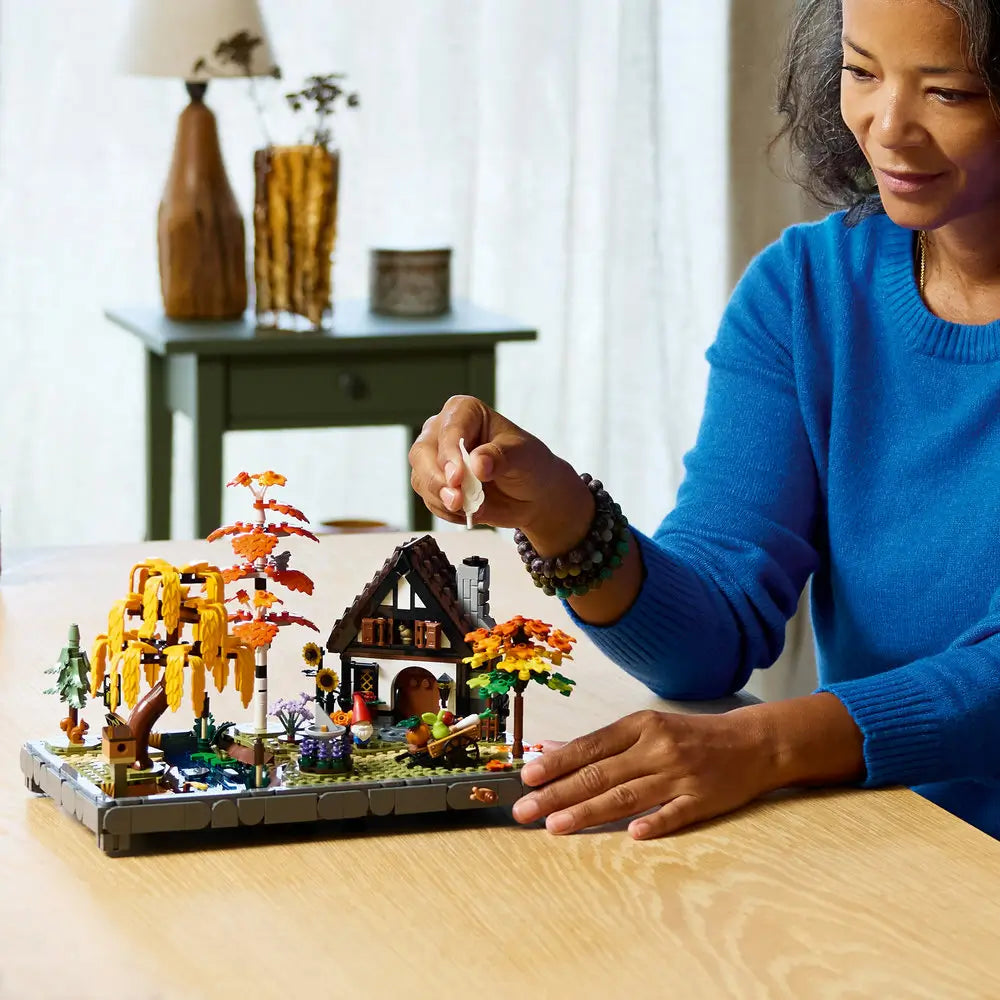 LEGO autumn cottage garden lifestyle image shows an adult carefully placing a small white chimney element onto the detailed cottage, demonstrating hands-on building and display enjoyment in a home setting.