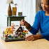 LEGO autumn cottage garden lifestyle image shows an adult carefully placing a small white chimney element onto the detailed cottage, demonstrating hands-on building and display enjoyment in a home setting.