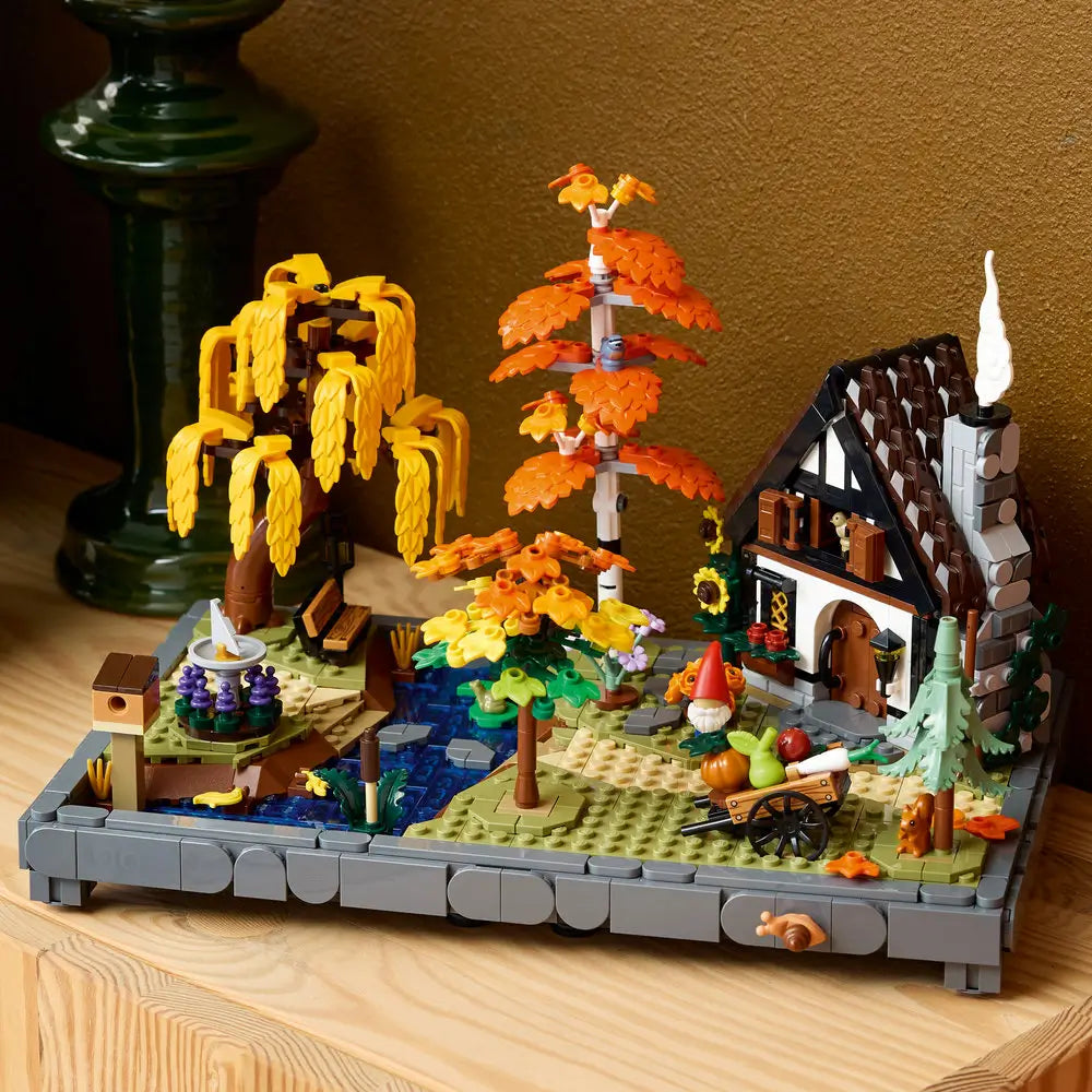 LEGO autumn cottage garden displayed shows orange and yellow trees, a stone cottage, water feature, and garden accessories built from plastic bricks, styled as a cozy decorative centerpiece indoors.