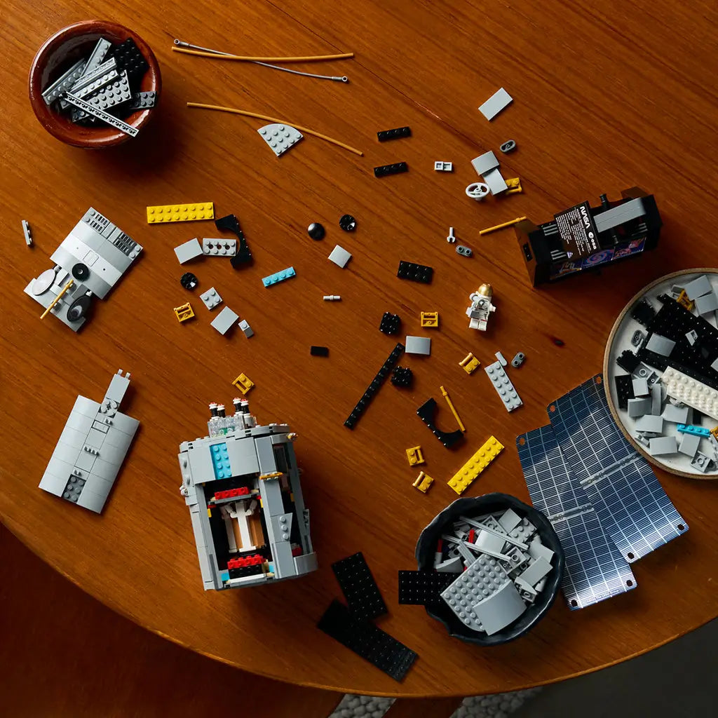 LEGO Icons Hubble Space Telescope set is shown partly assembled in gray, black and gold plastic bricks, with blue solar panels and loose pieces spread across a wood table.