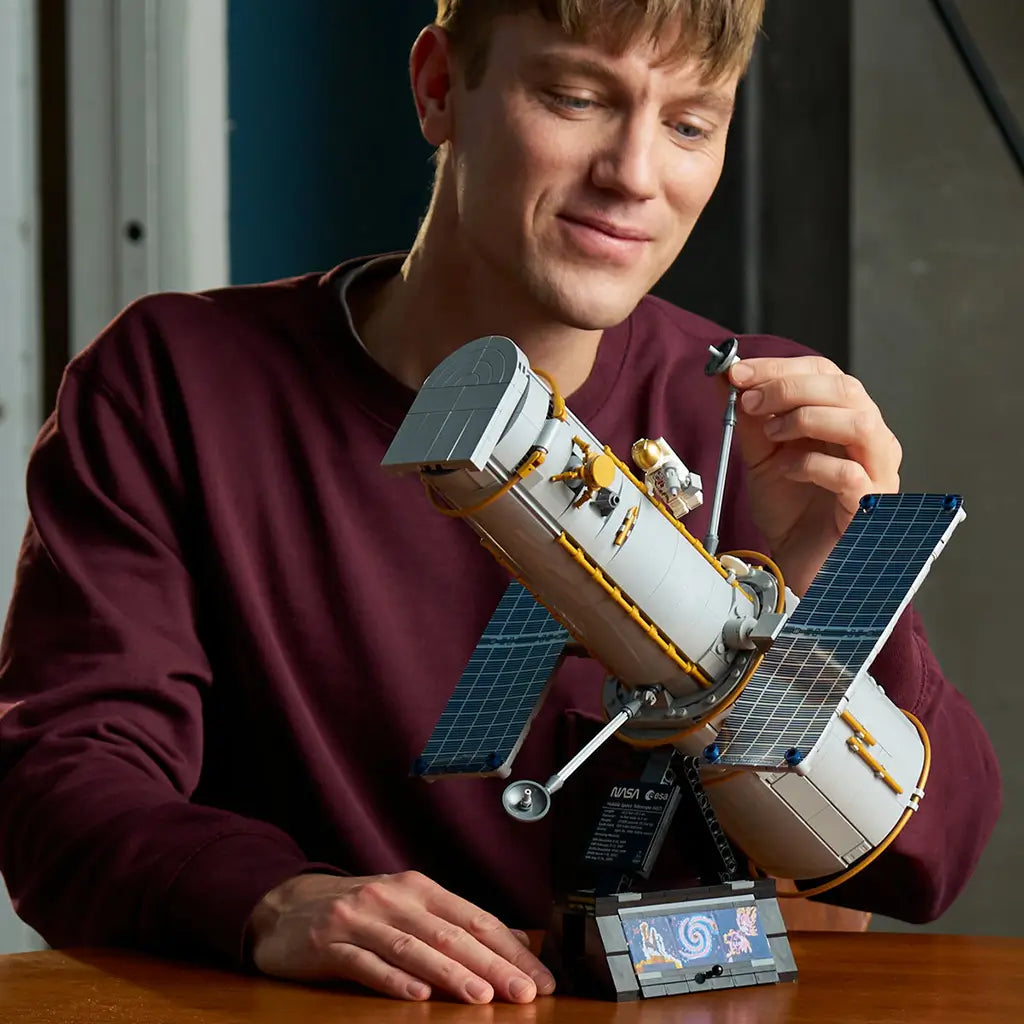 LEGO Icons Hubble Space Telescope model in gray, gold and blue is adjusted by an adult builder, with solar panels, astronaut figure and black display stand on a wood table.