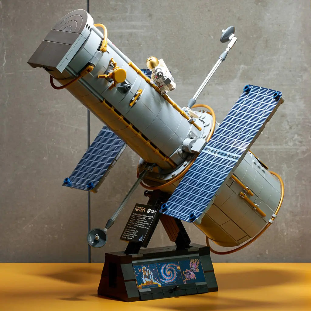 LEGO Icons Hubble Space Telescope model features a gray brick-built cylindrical body, gold details, blue solar panels and astronaut figure on a black display stand indoors.
