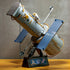 LEGO Icons Hubble Space Telescope model features a gray brick-built cylindrical body, gold details, blue solar panels and astronaut figure on a black display stand indoors.