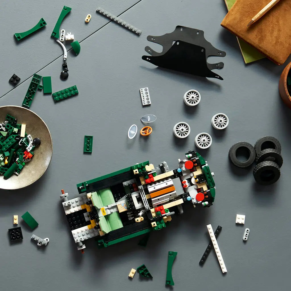 LEGO Icons Jaguar E-Type build is partly assembled from dark green, tan, black and gray plastic bricks, with loose wheels, tires and body panels arranged on a gray table.