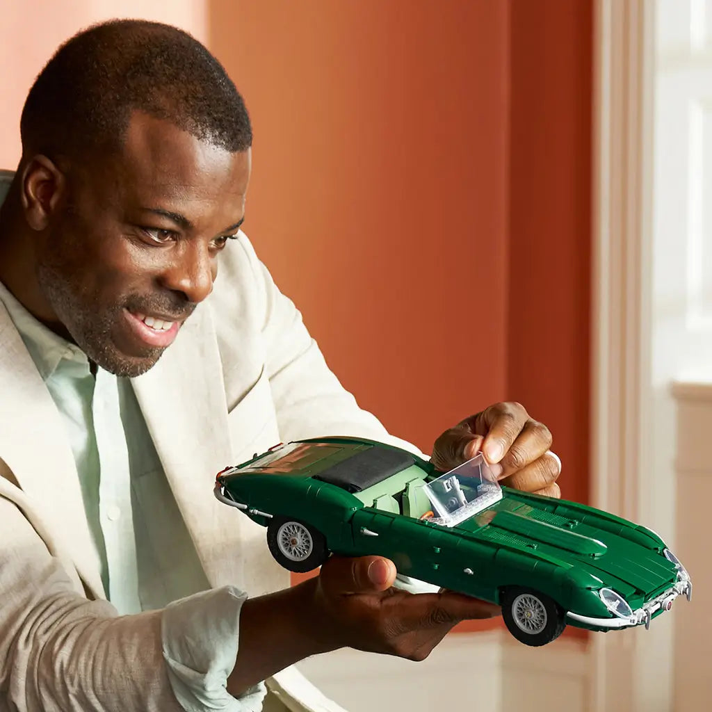 LEGO Icons Jaguar E-Type model in dark green plastic bricks is held by an adult builder, showing its long roadster body, clear windshield and silver wheels indoors.