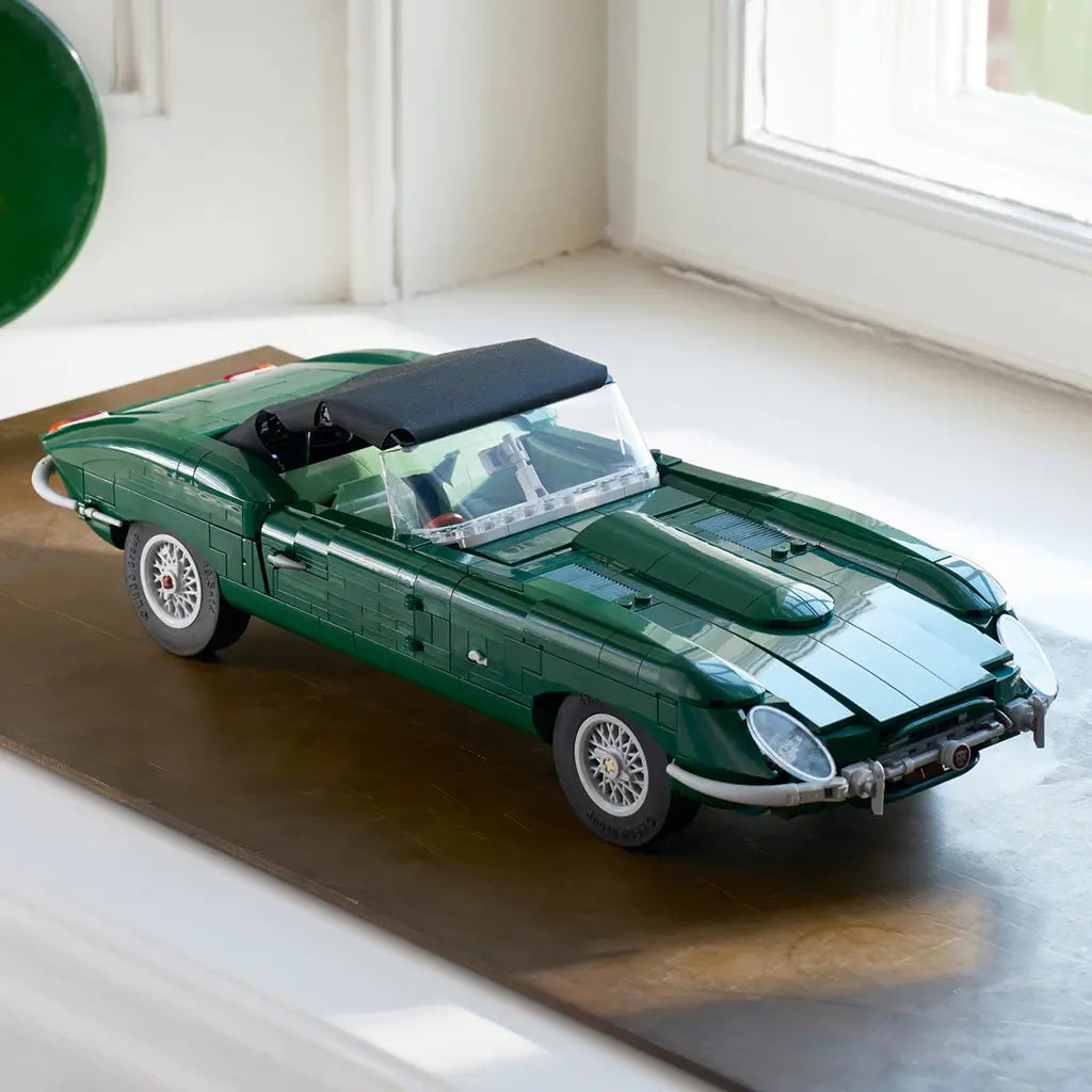 LEGO Icons Jaguar E-Type display features a dark green brick-built roadster with black soft top, clear windshield and silver wire wheels resting on a wood surface by a window.