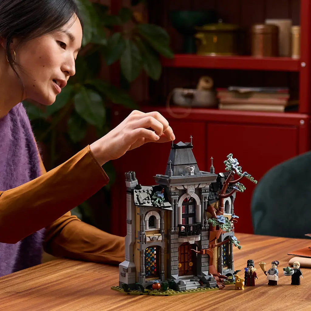 LEGO Icons Mayor Manor set 11383 lifestyle scene shows an adult adjusting the gray and brown plastic gothic mansion beside three minifigures and a cat on a wood table.
