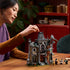 LEGO Icons Mayor Manor set 11383 lifestyle scene shows an adult adjusting the gray and brown plastic gothic mansion beside three minifigures and a cat on a wood table.
