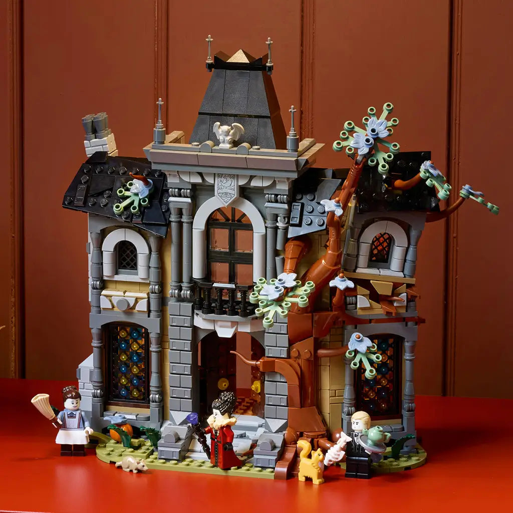 LEGO Icons Mayor Manor set 11383 displays a gray and brown plastic gothic mansion with stained-glass windows, twisting tree, three minifigures and animals on a red tabletop.