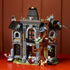 LEGO Icons Mayor Manor set 11383 displays a gray and brown plastic gothic mansion with stained-glass windows, twisting tree, three minifigures and animals on a red tabletop.